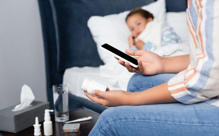 Mother holding bottle of pills and mobile phone near sick daughter lying in bed